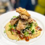 Crispy fried chicken perched on a thin tortilla with greens, sesame seeds, and a red sauce drizzle on a white plate.